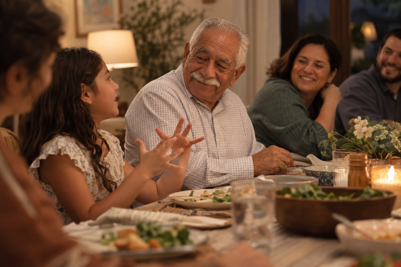 A grandfather grinning as his granddaughter tells him a story at a family dinner