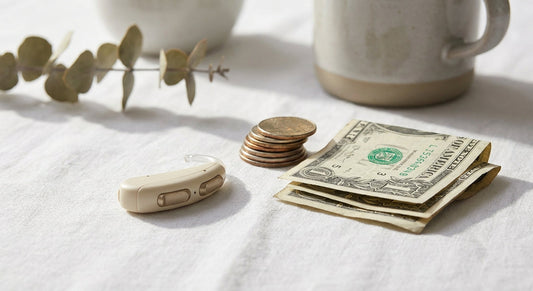 A modern hearing aid resting beside a small stack of coins and folded dollar bills on a clean white surface, lit with soft natural light.